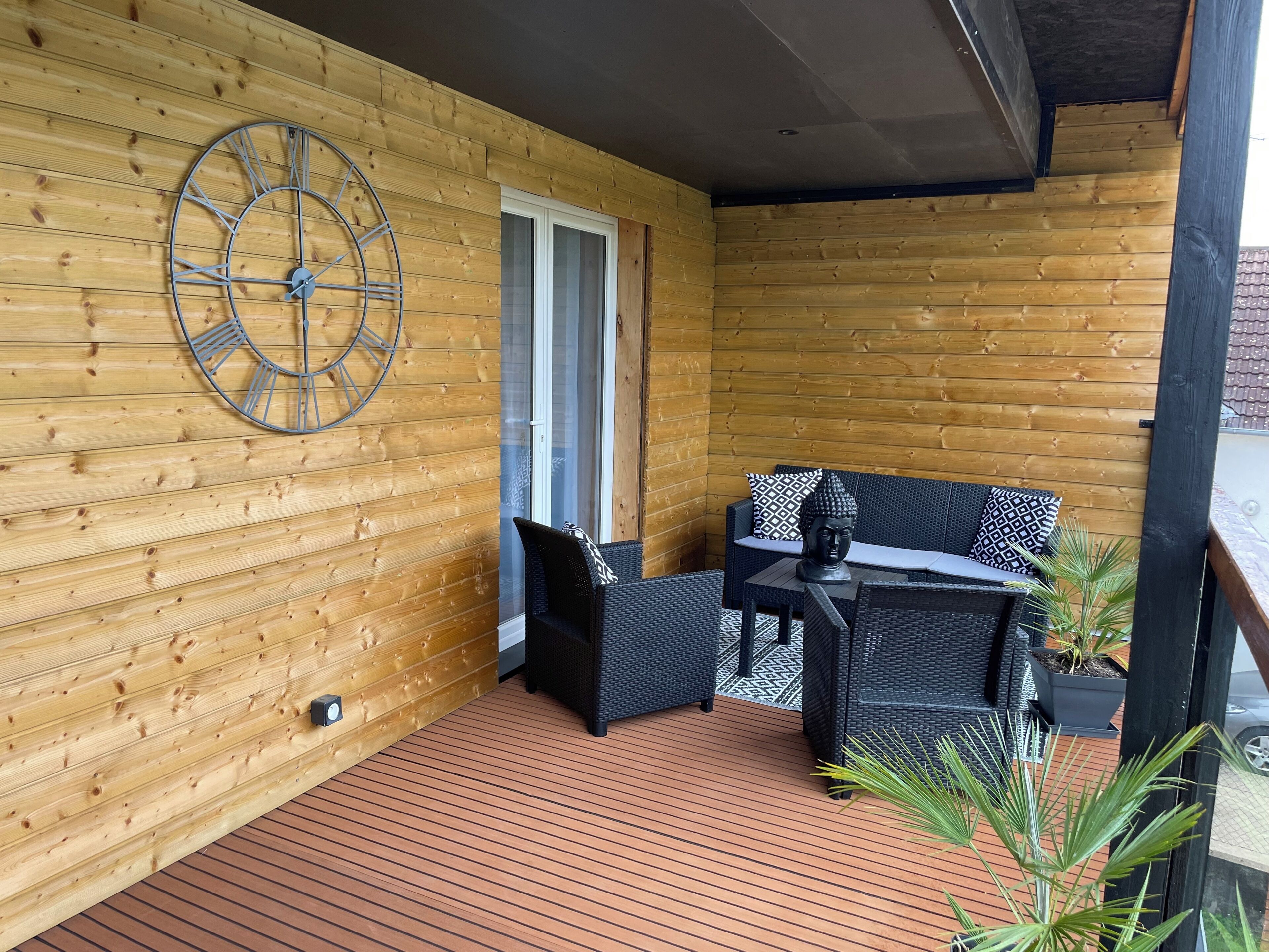 Una terraza de madera con una mesa y sillas de mimbre negro, un reloj de pared grande y una puerta que da a un espacio interior.