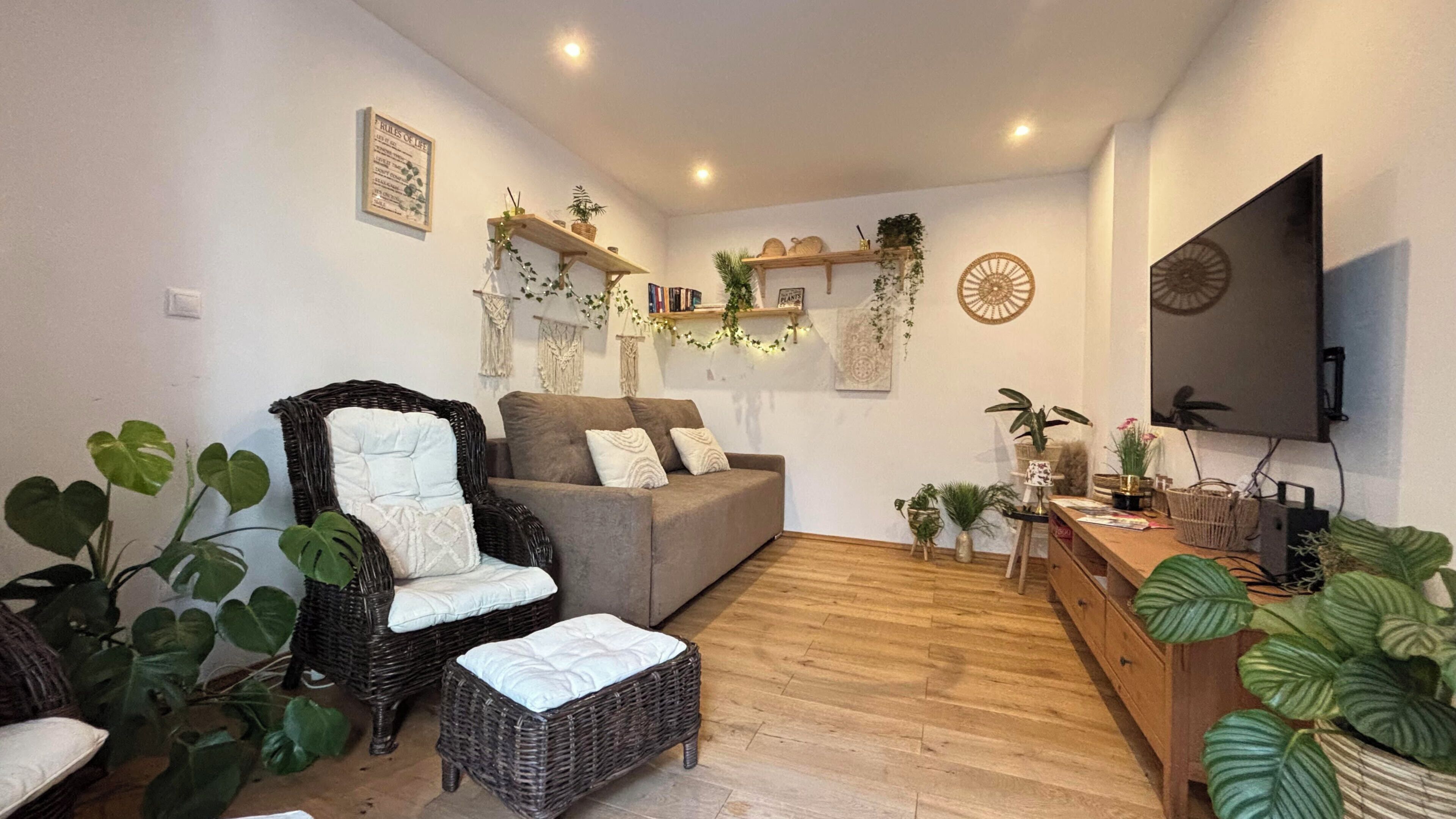 Un salon comprenant un canapé, un fauteuil en osier, un petit pouf et diverses plantes en pot.
