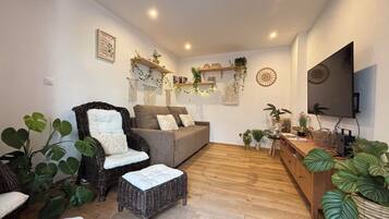 Un salon comprenant un canapé, un fauteuil en osier, un petit pouf et diverses plantes en pot.
