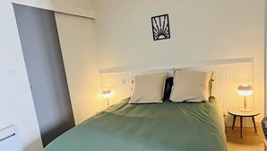 A bedroom with a bed, two bedside lamps, a wall-mounted light fixture, and a radiator. - "Sauge de Sarrail" City Center Apartment (Saintes)