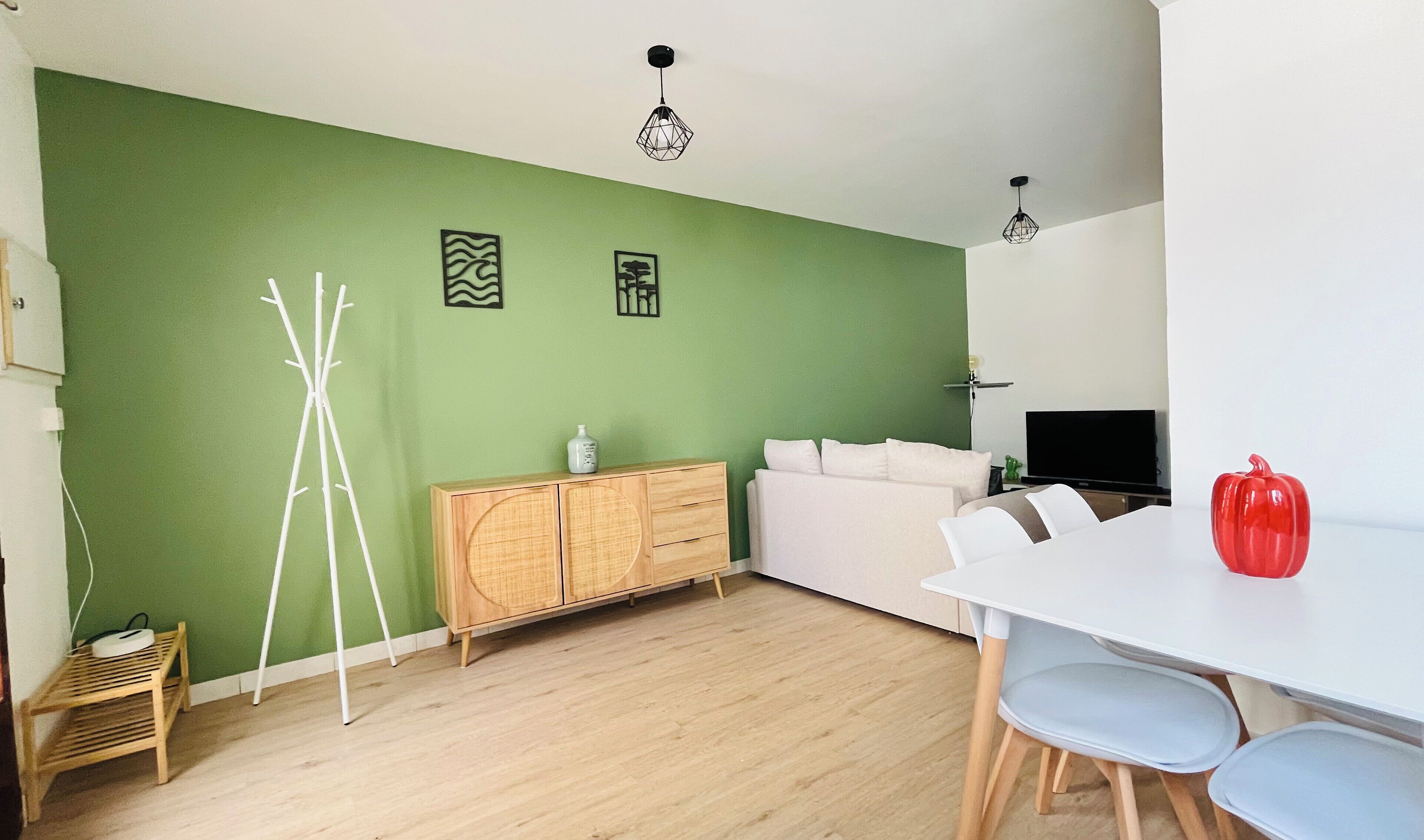 A room with a green wall, a white sofa, a dining table with chairs, a wooden cabinet, and a red object on the table.
