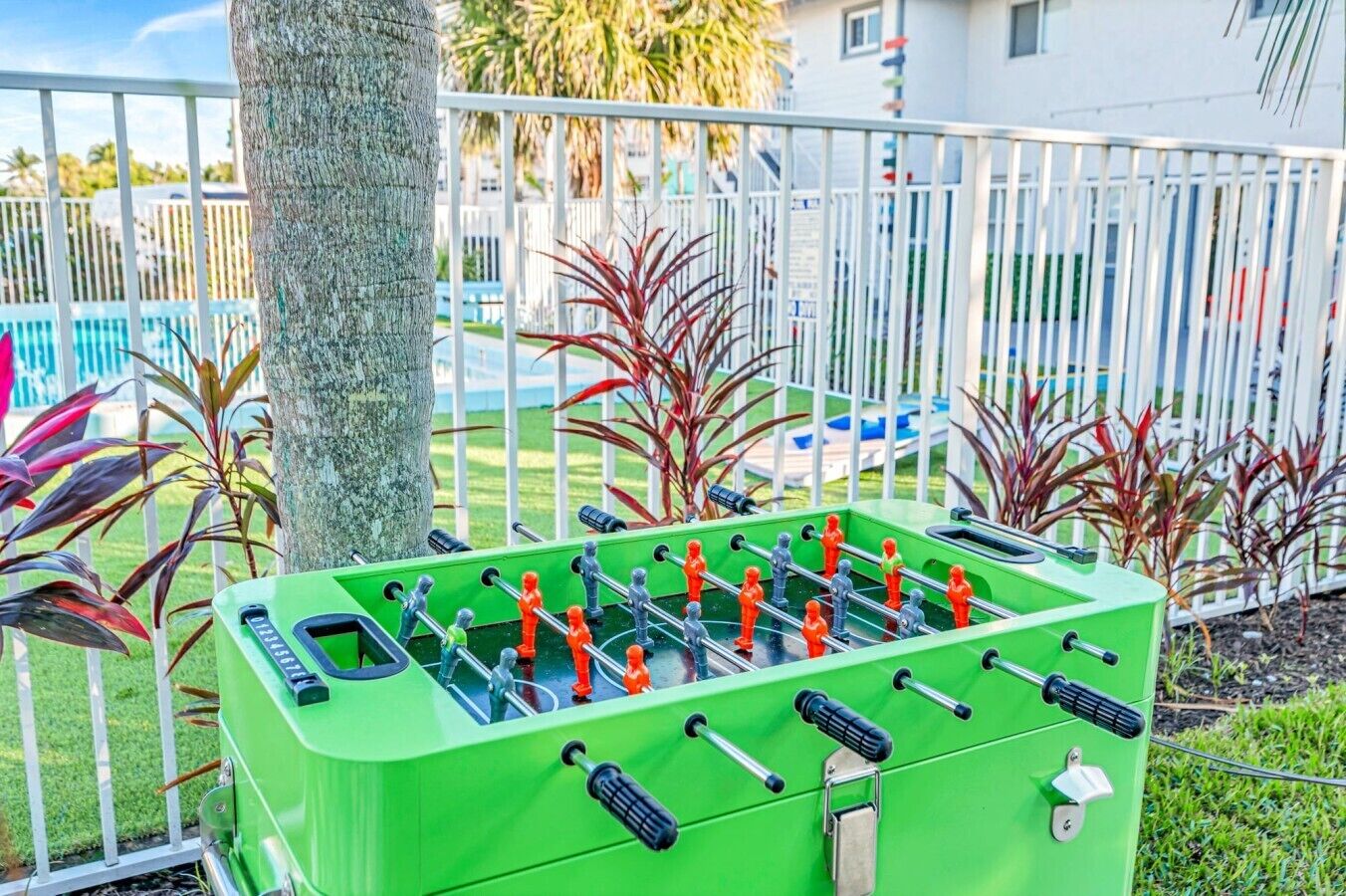A green foosball table with a metal railing and plants in the background.