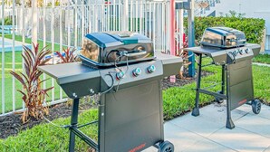Twee draagbare gasgrills met wielen, merk 'megamaster', staan buiten op een geplaveid terras, met een witte hek en begroeiing op de achtergrond.