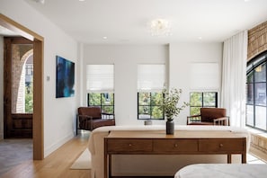A spacious bedroom with a large bed, two chairs, a desk, and a view of greenery through windows. - Waterfront Luxury Home Downtown Traverse City (Traverse City)