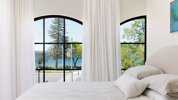 A bedroom with a large bed, white bedding, and a view of trees and a body of water through a window. - Waterfront Luxury Home Downtown Traverse City (Traverse City)