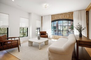A modern living room with a sofa, armchair, and coffee table, large windows with blinds, a brick fireplace, and a view of the outside. - Waterfront Luxury Home Downtown Traverse City (Traverse City)