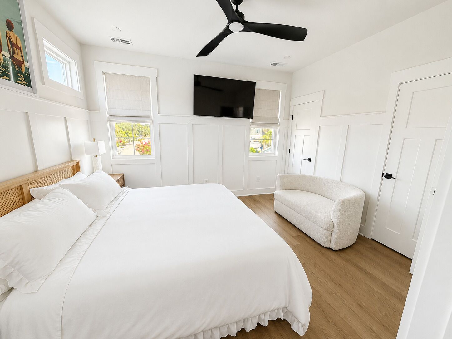 A bedroom with a bed, a white armchair, a ceiling fan, a flat-screen TV, and a window with a view of greenery.