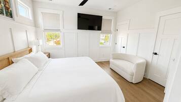 A bedroom with a bed, a white armchair, a ceiling fan, a flat-screen TV, and a window with a view of greenery.