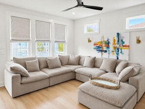 Una sala moderna con un sofá seccional color beige, un ventilador de techo y un cuadro abstracto grande en la pared.