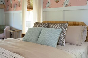Ein ordentlich gemachtes Bett mit einer beigen Tagesdecke und zwei Kissen, ein Nachttisch mit einer Lampe und ein Sessel in einem Zimmer mit Blumenmuster-Tapete.