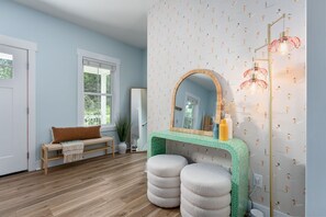 Ein Zimmer mit einer hölzernen Bank, einem runden Spiegel, einem grün gemusterten Konsolentisch und einer Stehlampe.