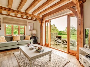 A cozy living room with a sofa, coffee table, and a fireplace, featuring wooden beams and large windows. - Luxury Cotswolds Cottage for 7 (Tewkesbury)