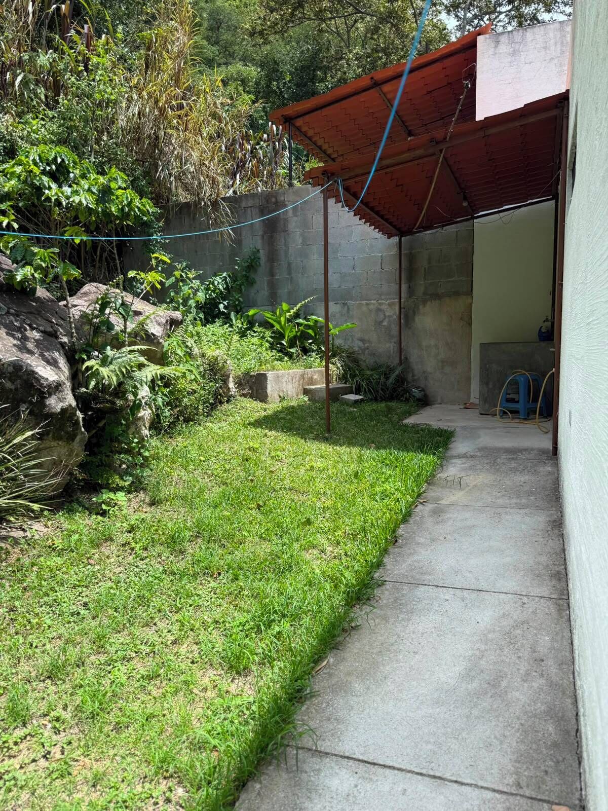 Un sendero de concreto que conduce a un área techada con techo rojo, rodeada de follaje y un muro de piedra.