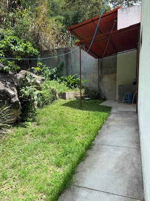 Un sendero de concreto que conduce a un área techada con techo rojo, rodeada de follaje y un muro de piedra.