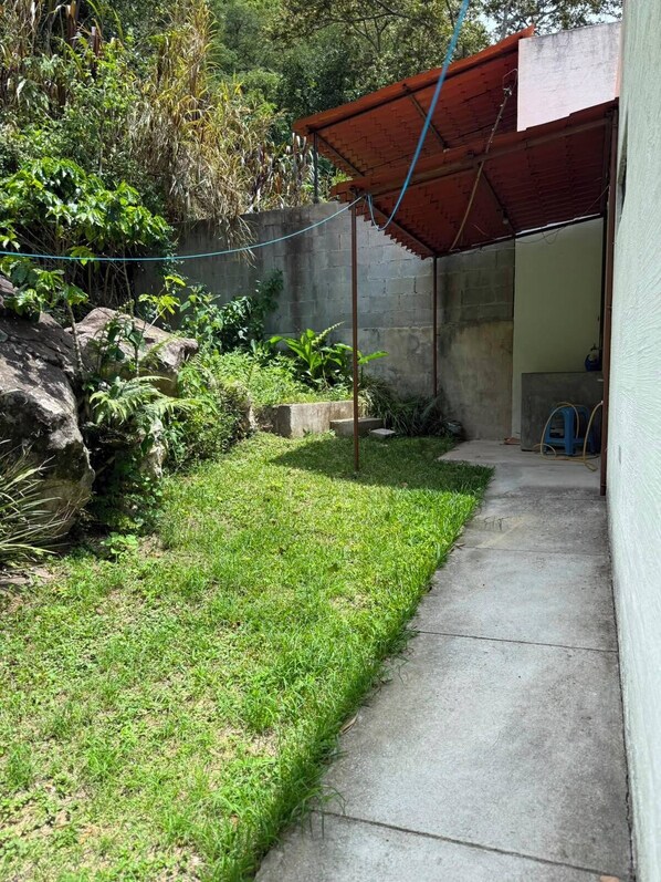 A concrete pathway leading to a covered area with a red roof, surrounded by greenery and a stone wall.
