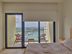 Un dormitorio con un gran ventanal corredera que da a un balcón con vistas a un puerto deportivo y edificios.