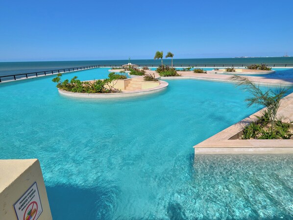 A large, clear blue pool with a circular island and palm trees, surrounded by a black railing. - Luxury Oceanfront Condo | Marina Resort | 3BR | Pools | Sleeps 10 (Progreso)