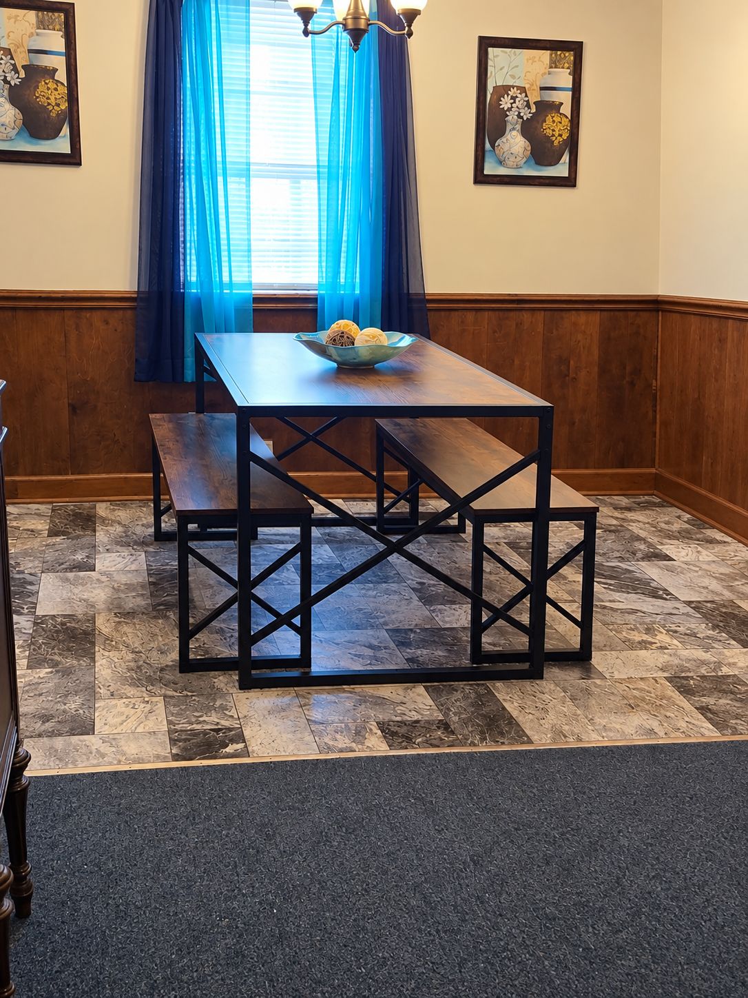 Un coin repas avec une table et des bancs en bois, un rideau bleu et deux tableaux encadrés accrochés au mur.