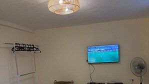 Quarto com cama, televisão na parede, ventilador, penteadeira e varal para roupas.