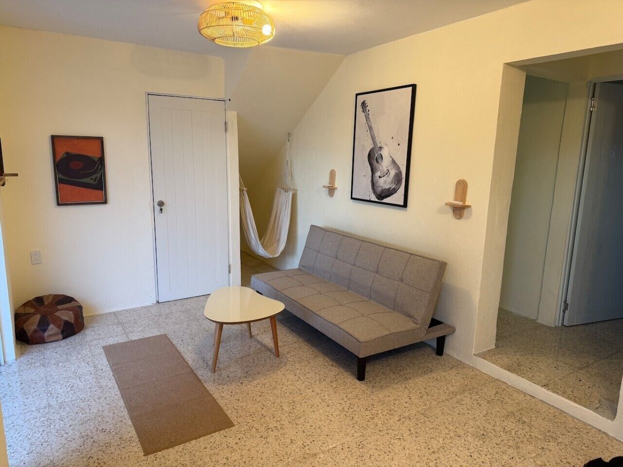 Una habitación con sofá, una mesita, una hamaca, una puerta y una pared con un cuadro de guitarra.