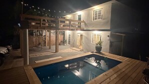 Uma piscina com deck de madeira, uma casa com várias janelas e uma área externa coberta.