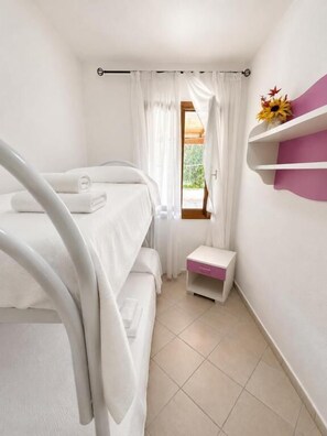 A single bed with white bedding, a small bedside table, a window with curtains, and a shelf with a vase of flowers. - Beach Walk Apartments - San Teodoro (San Teodoro)