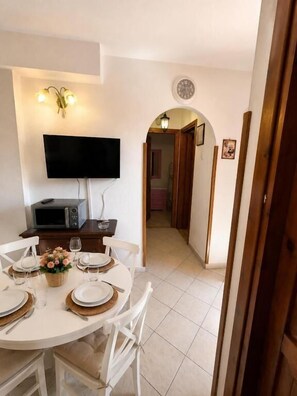A dining area with a round table, white chairs, a flat-screen TV, a microwave, and a small table with a vase of flowers. - Beach Walk Apartments - San Teodoro (San Teodoro)