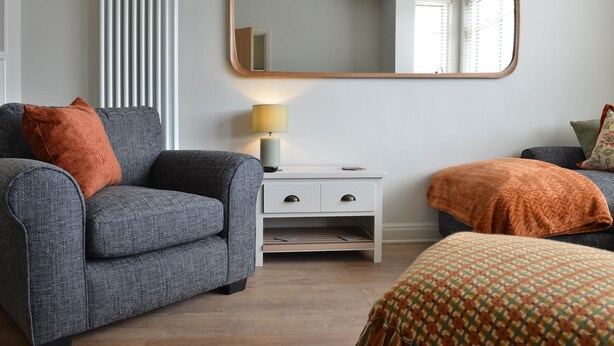 Una habitación con un sofá gris, una mesita de noche blanca con cajones, un espejo y un otoman con estampado.