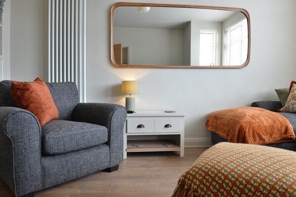 A room with a grey sofa, a white nightstand with drawers, a mirror, and a patterned ottoman.