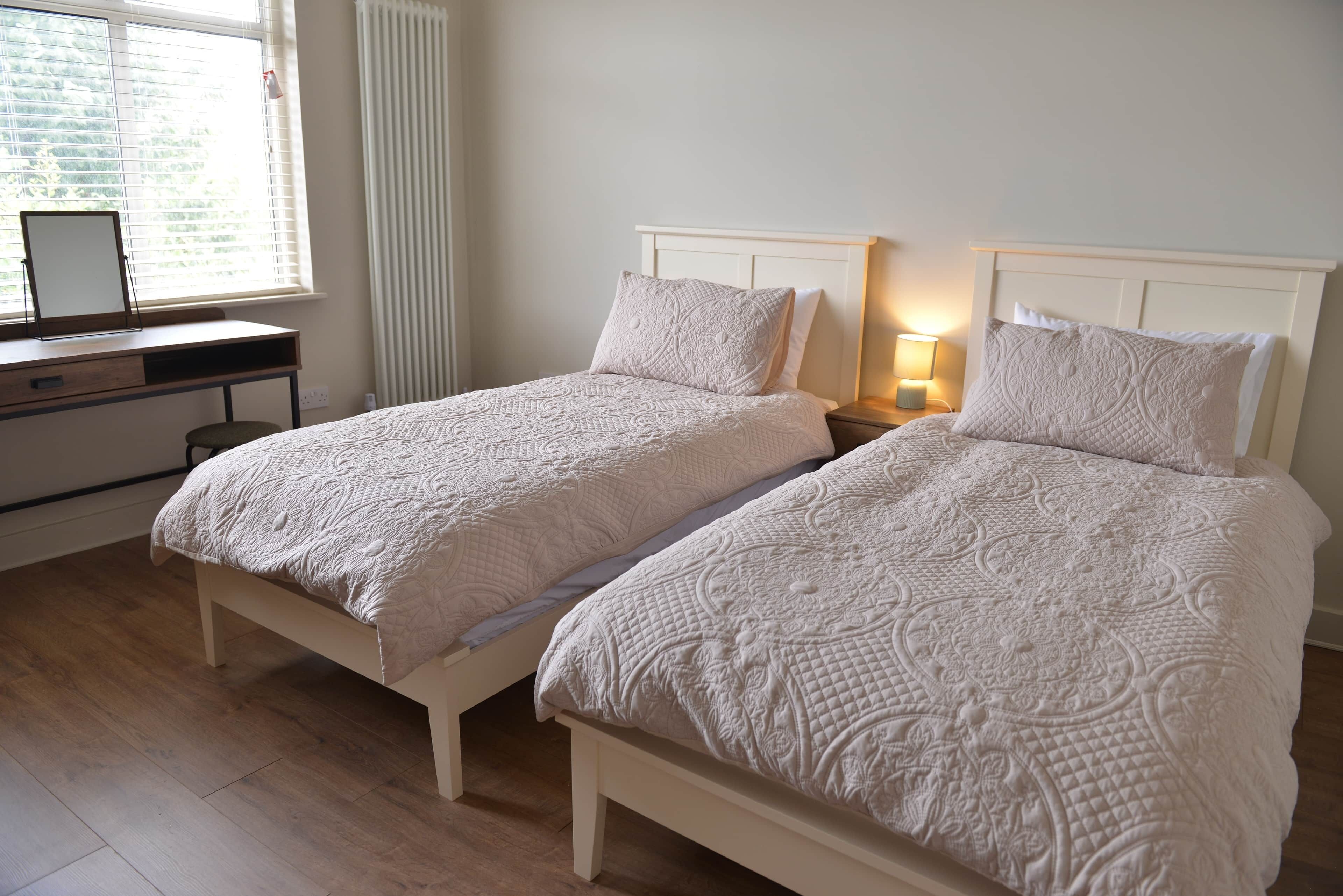 Une chambre avec deux lits simples, un bureau et une fenêtre avec des stores.