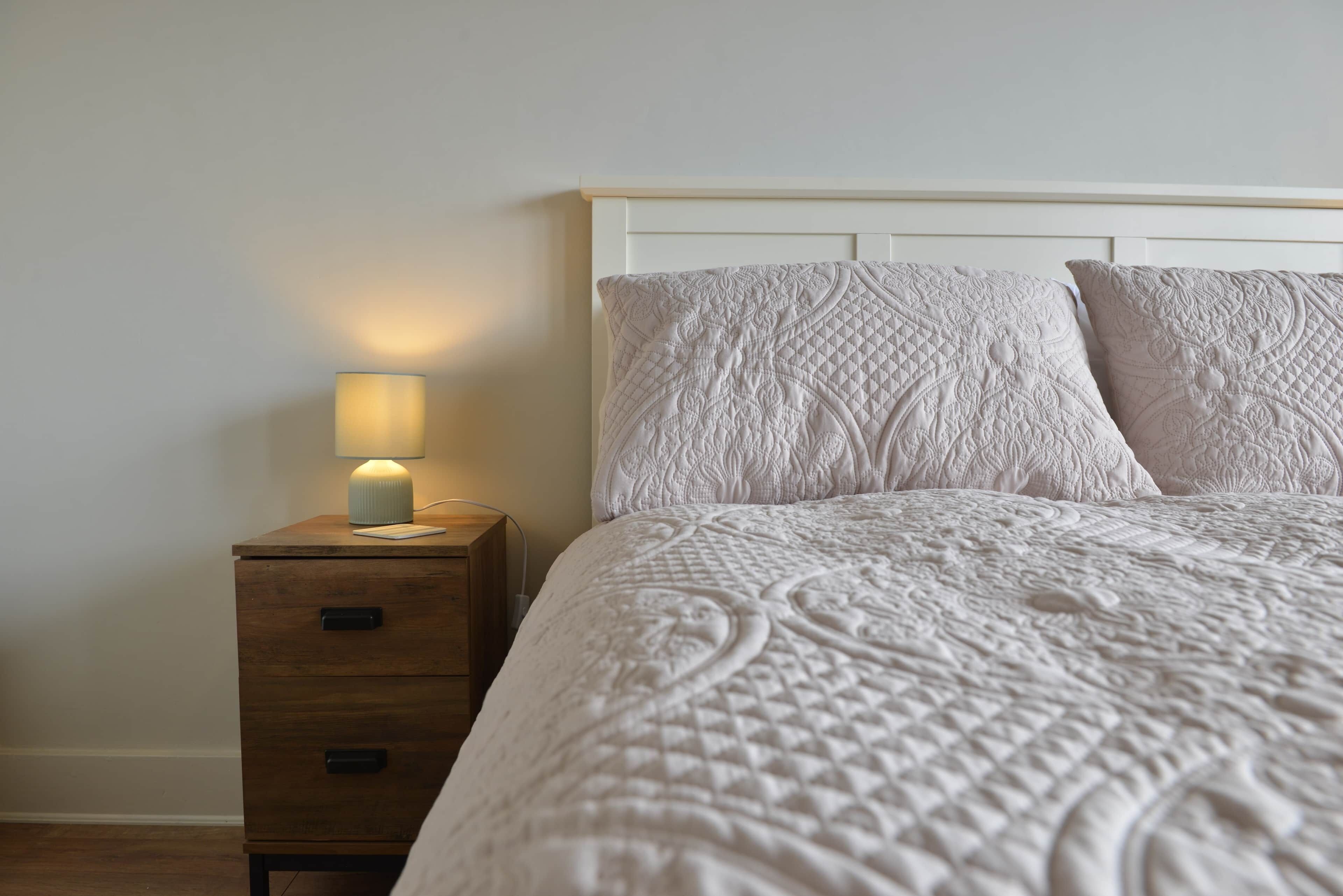 A neatly made bed with a quilted headboard and pillows, a bedside table with a lamp, and a wooden nightstand with drawers.