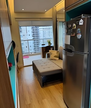 Ein kompakter Wohnraum mit Küchenzeile, einem Bett, einem kleinen Tisch und Blick auf eine Stadtlandschaft durch ein großes Fenster.