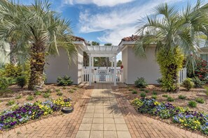 A gated entrance with a white pergola, palm trees, and flower beds. - Shore Beat Works - Emerald Water (Miramar Beach)