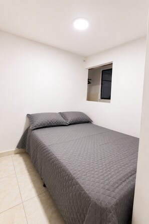 Ein Einzelbett mit gesteppter, grauer Bettdecke, ein kleines Fenster mit schwarzem Rahmen und eine weiße Wand.