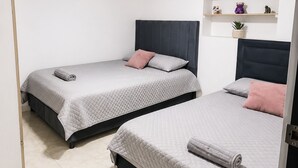 Duas camas de solteiro com cabeceiras, uma parede branca, uma prateleira com objetos decorativos e uma porta.