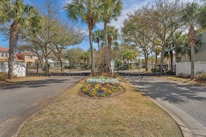 A gated residential area with palm trees, a flower bed, and a paved road. - Ahoy Matey - Emerald Waters (Miramar Beach)