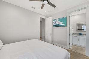 A bedroom with a bed, a ceiling fan, a painting, and a bathroom with a mirror and cabinets. - Ahoy Matey - Emerald Waters (Miramar Beach)