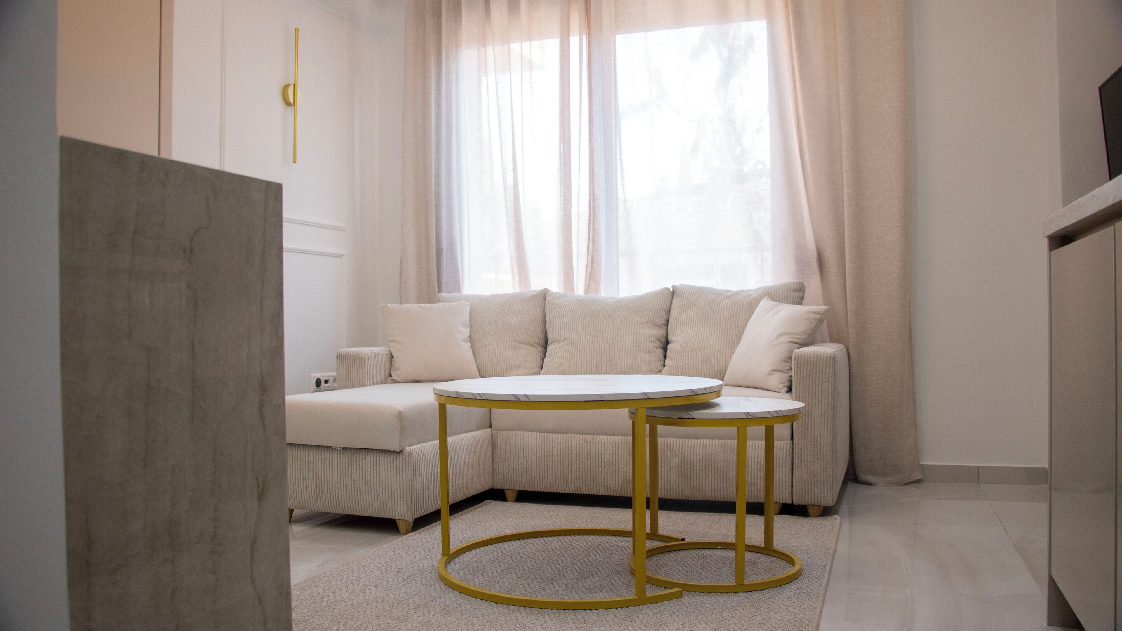 Una sala moderna con un sofá color beige, una mesa redonda con marco dorado y un ventanal con cortinas translúcidas.