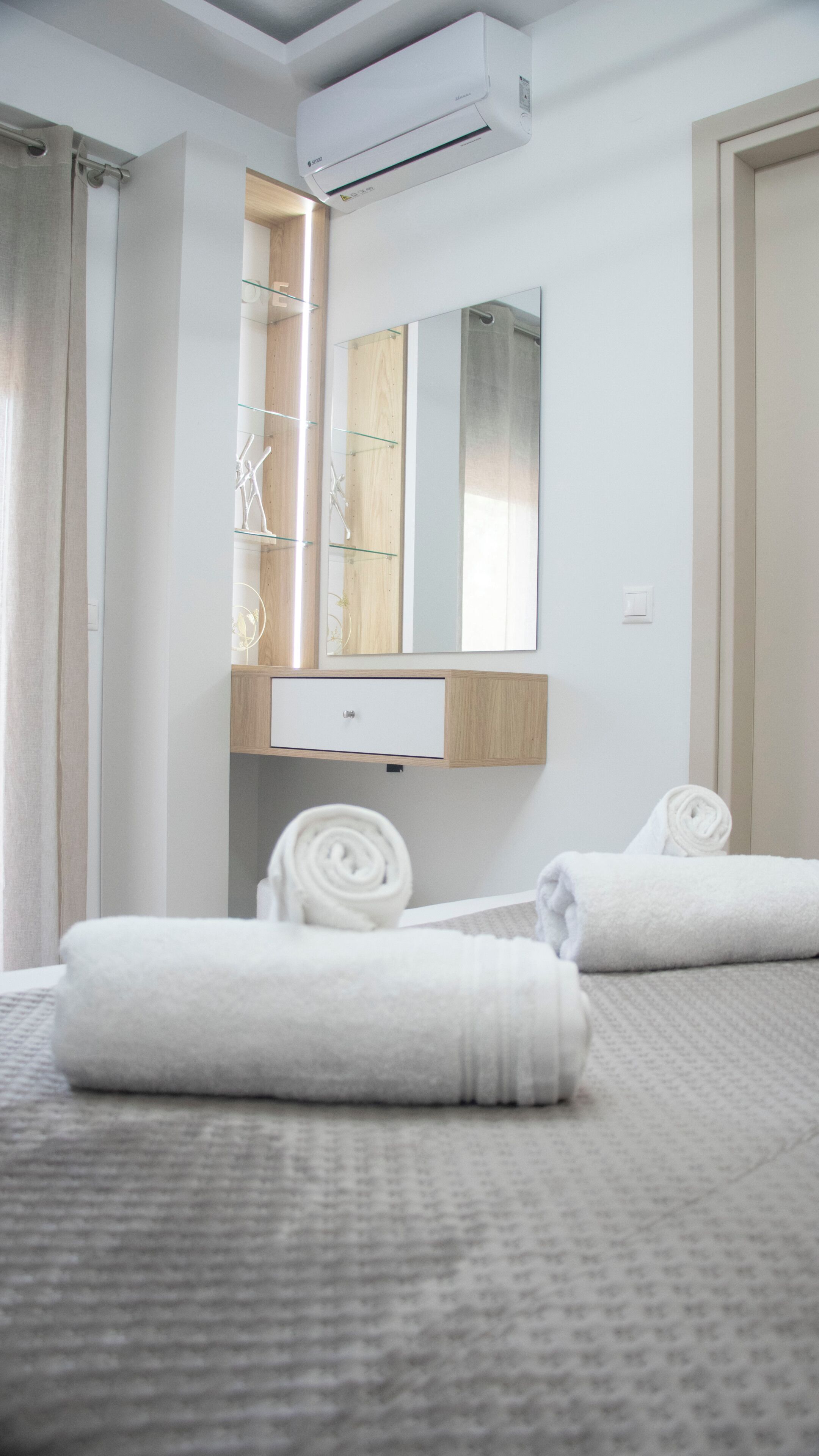 Habitación de hotel con cama, dos toallas enrolladas, un espejo, un aparador de madera y un equipo de aire acondicionado.