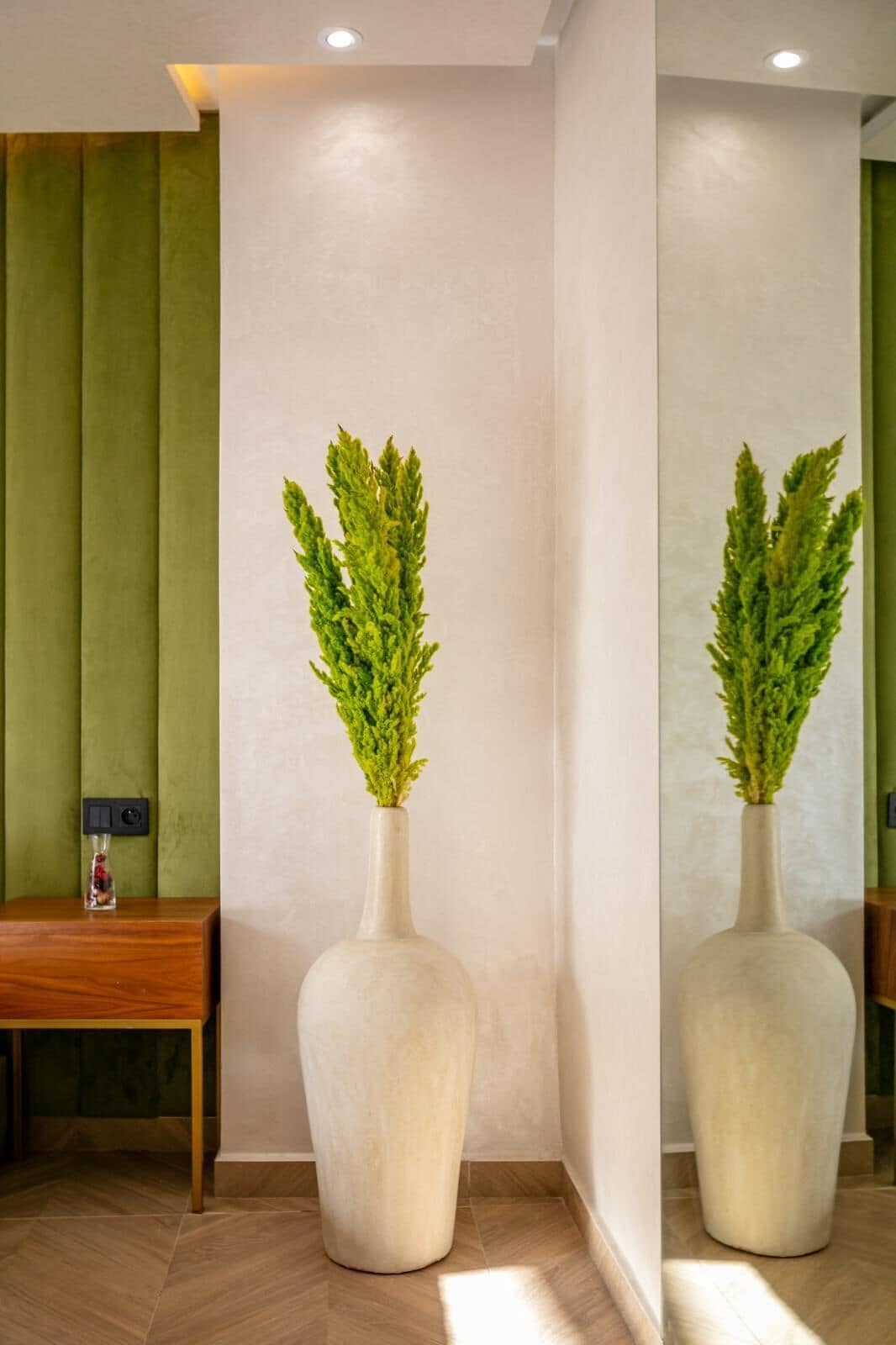 A room with a large mirror, a green accent wall, a wooden table, and a white vase with green foliage.