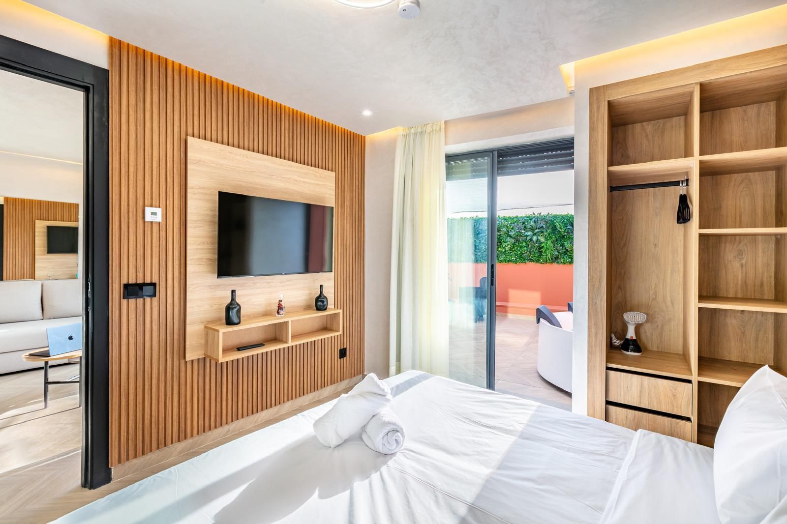 Une chambre d’hôtel moderne équipée d’un lit, d’un téléviseur fixé sur un mur recouvert de panneaux en bois, d’une porte vitrée donnant sur un balcon et d’une armoire en bois.