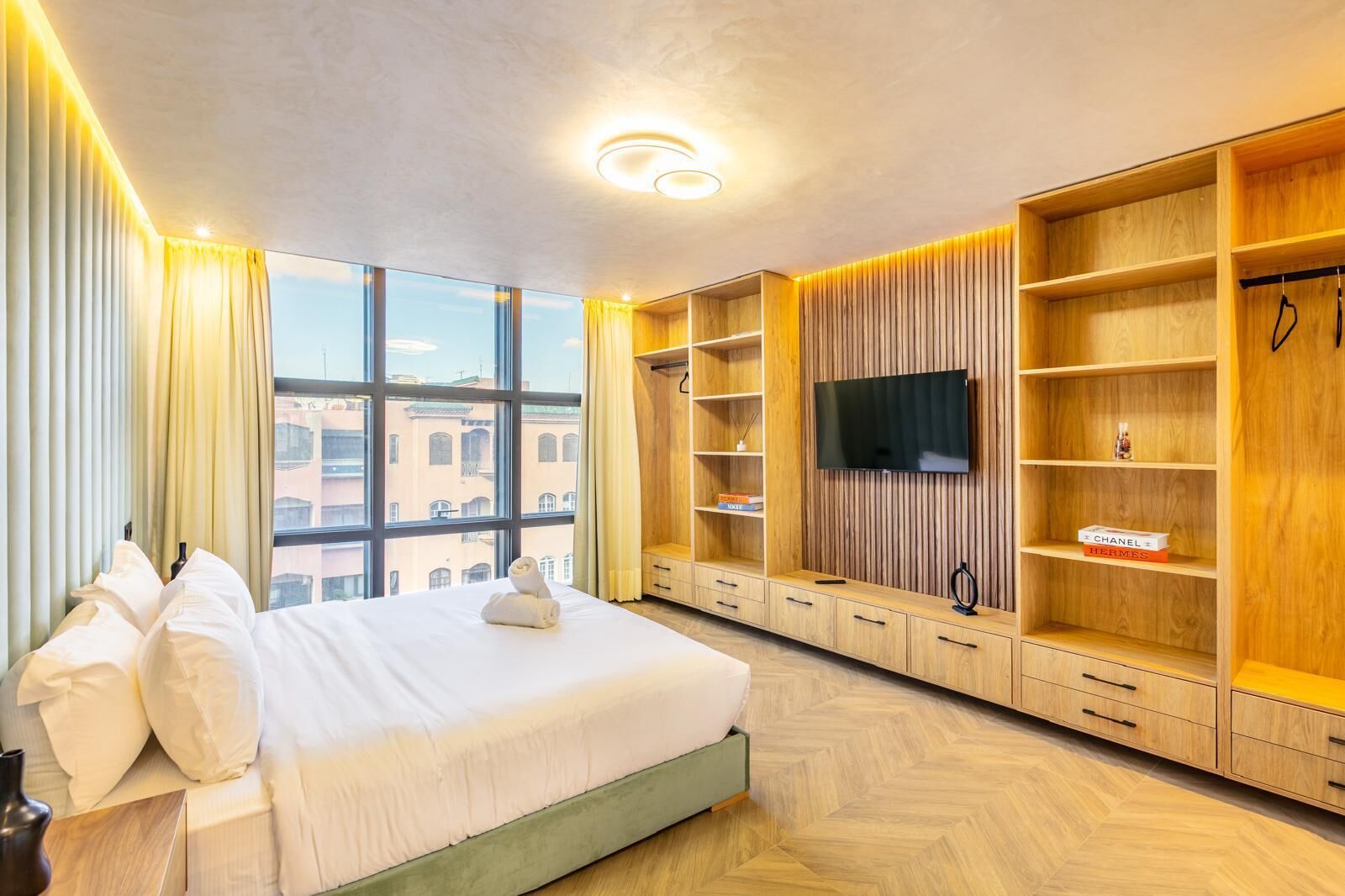 A hotel room with a large bed, a television, wooden shelving, and a view of buildings through large windows.