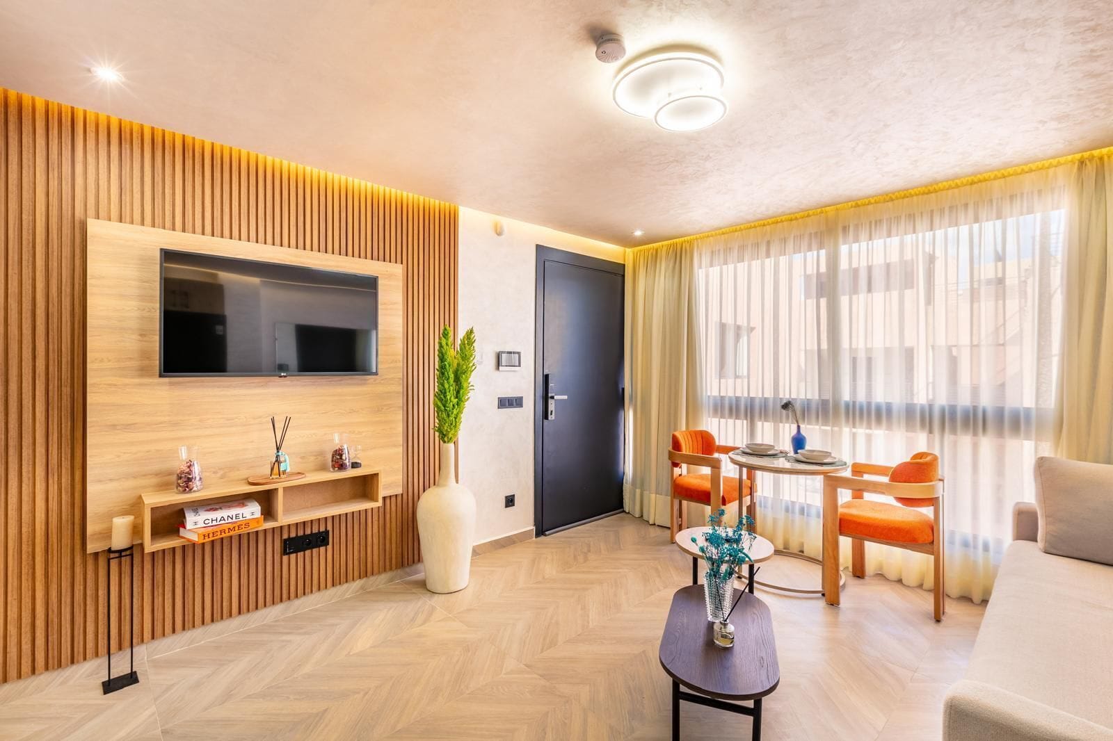 A modern living room with a flat-screen TV, wooden paneling, a dining area with orange chairs, and a large window with sheer curtains.