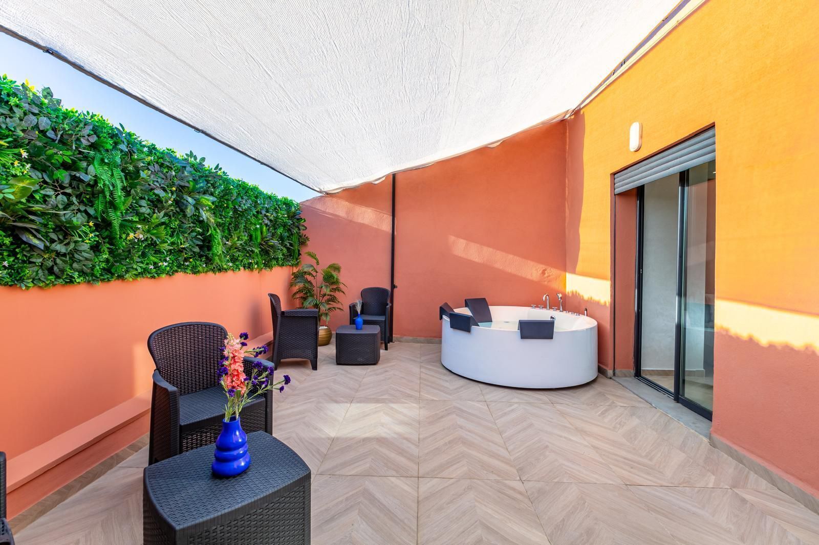 A sunlit outdoor seating area with a round hot tub, wicker furniture, and a wall of greenery.
