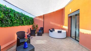A sunlit outdoor seating area with a round hot tub, wicker furniture, and a wall of greenery.