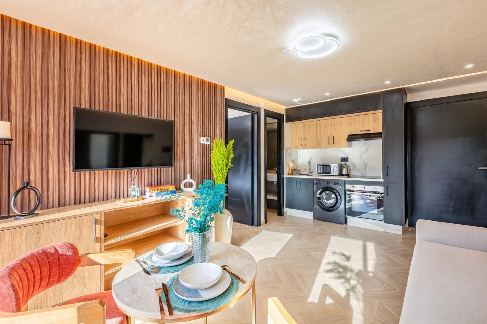 A modern living room with a flat-screen TV, wooden panel wall, dining table with plates, and a sofa.