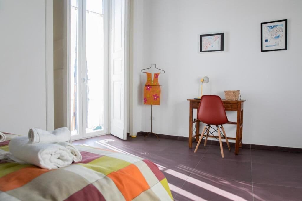 A bedroom with a bed, a desk, and a chair. There is a window with curtains, a map on the wall, and a towel on the bed.