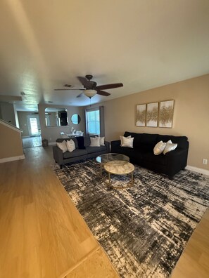 Ein geräumiges Wohnzimmer mit einem dunklen Sofa, einem runden Couchtisch und einem gemusterten Teppich. Ein Deckenventilator ist sichtbar, und an der Wand hängen gerahmte Bilder.