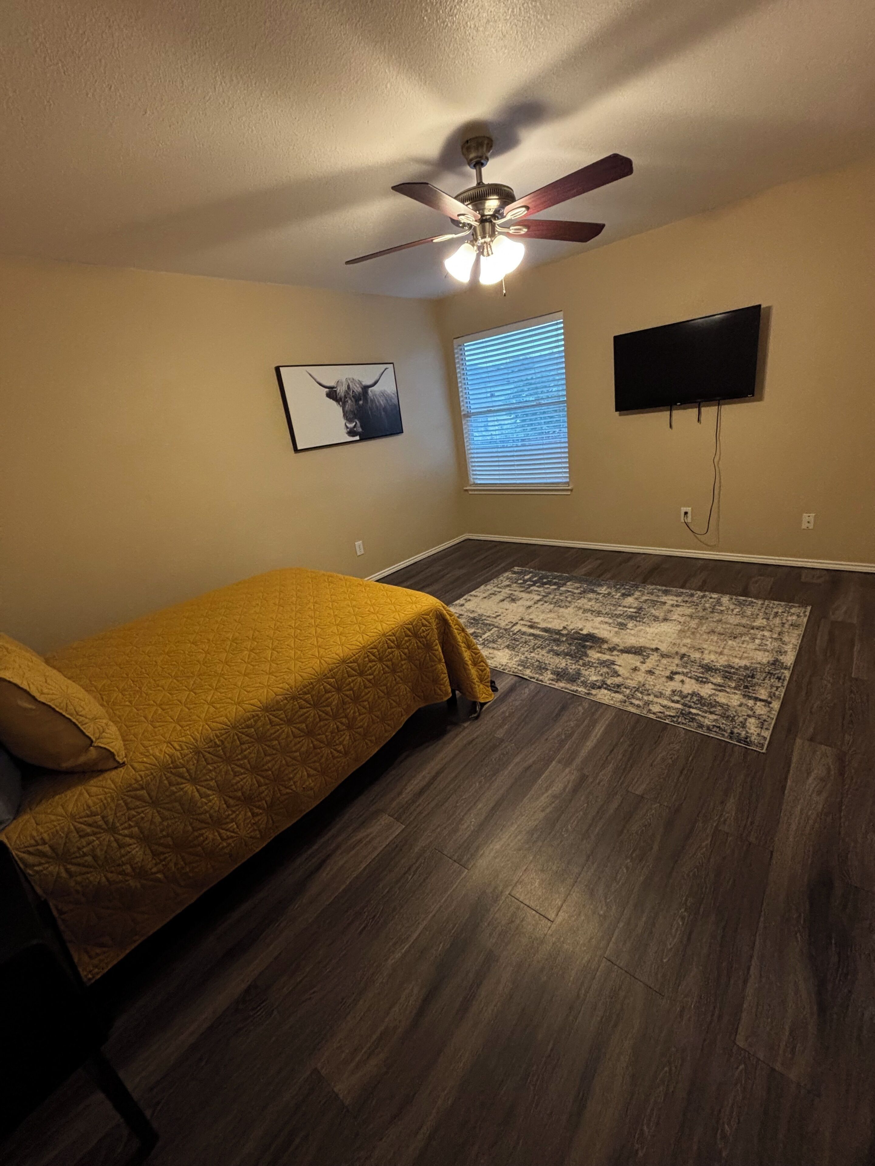 Un dormitorio con cama, ventilador de techo, televisor montado y ventana con persianas.