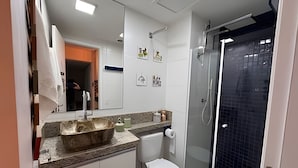 A bathroom with a white toilet, a sink with a stone basin, a glass shower enclosure, and a mirror with artwork. - Confortável Studio na Bela Vista Centro (São Paulo)